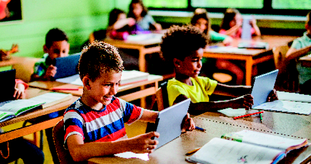 Imagem: Fotografia. Uma sala de aula com as carteiras de madeira organizadas em duplas. No primeiro plano, dois alunos segurando tablets: um menino loiro de camiseta branca, azul e vermelha listrada e uma menina negra de camiseta amarela. Ao fundo, mais alunos nas carteiras.  Fim da imagem.