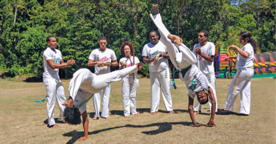 Imagem: Fotografia. Dois homens negros usando calça e camiseta branca lutando capoeira. Ambos estão com os braços apoiados no chão e as pernas para cima. Atrás, cinco pessoas de roupa branca olham e batem palma.  Fim da imagem.