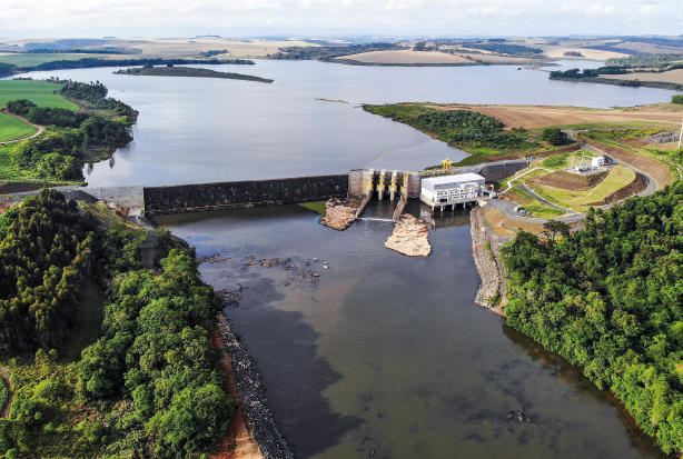 Imagem: Fotografia. Uma barragem com uma usina de energia do lado. Ao redor, árvores.  Fim da imagem.