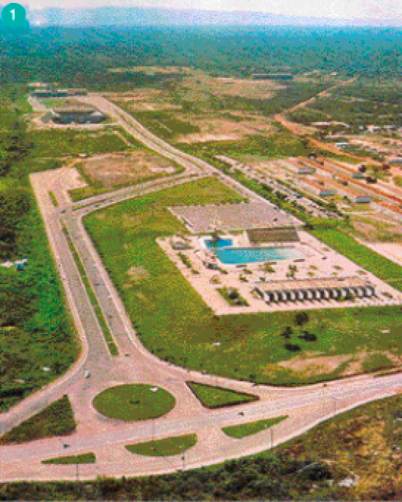 Imagem: Fotografia. Uma rodovia em construção, com uma rotatória na parte da frente e ao lado algumas construções espaçadas, uma delas com uma piscina.  Fim da imagem.