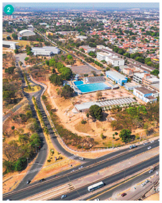 Imagem: Fotografia. Uma rodovia com uma construção com piscina ao lado e para a direita e no fundo, uma cidade.  Fim da imagem.