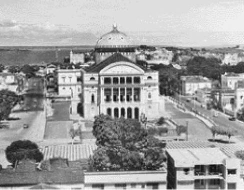 Imagem: Fotografia. Imagem em preto e branco e uma construção antiga com três andares, com pilares na frente e uma cúpula atrás. Na frente, uma praça com árvores.  Fim da imagem.