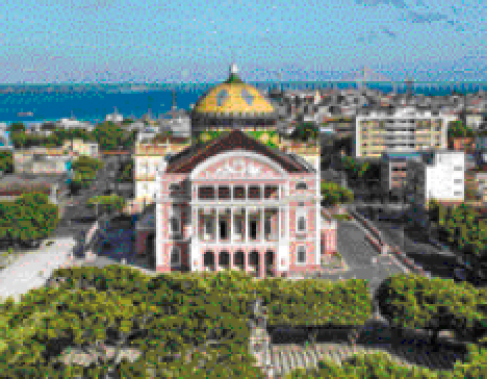 Imagem: Fotografia. Uma construção antiga com três andares, com pilares na frente e uma cúpula atrás. Na frente, uma praça com árvores. Ao fundo, mais construções e o mar.  Fim da imagem.