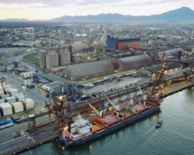 Imagem: Fotografia. Um navio atracado em um porto. Ao lado, construções e reservatórios cilíndricos. No fundo da imagem, uma cidade e montanhas.  Fim da imagem.