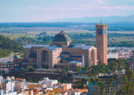 Imagem: Fotografia. Uma igreja com uma cúpula no meio e uma torre do lado direito. Ao redor dela há áreas de vegetação e na frente algumas casas e prédios menores.  Fim da imagem.