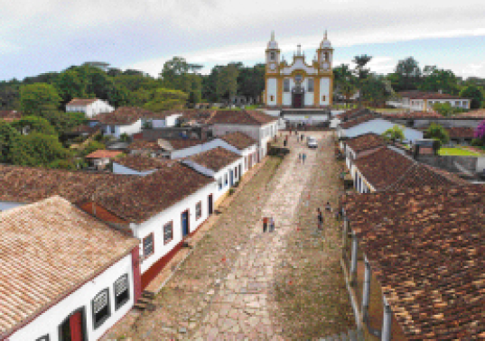 Imagem: Fotografia. Uma rua de pedra com algumas pessoas andando. Aos lados, casas antigas e no fundo uma igreja branca e amarela.  Fim da imagem.