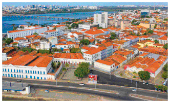 Imagem: Fotografia. Imagem aérea de uma cidade com casas com telhado de barro, uma avenida na frente e do lado esquerdo um rio com uma ponte.  Fim da imagem.