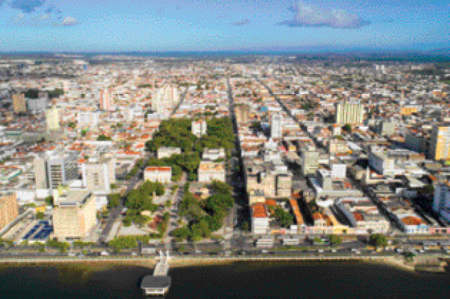 Imagem: Fotografia. Imagem aérea de uma cidade com ruas paralelas, formando quadras com casas e alguns prédios, e uma praia na frente.  Fim da imagem.