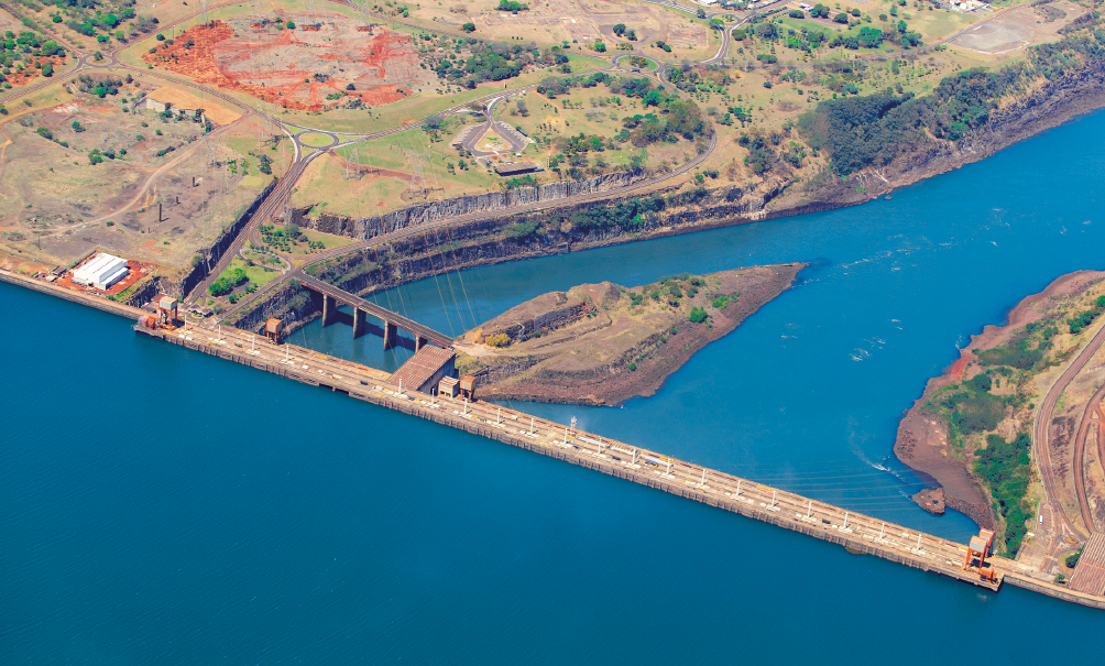 Imagem: Fotografia. Uma grande barragem com a construção de uma hidrelétrica do lado. Fim da imagem.