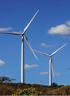 Imagem: Fotografia. Duas torres com hélices na parte de cima, para produção de energia eólica.  Fim da imagem.