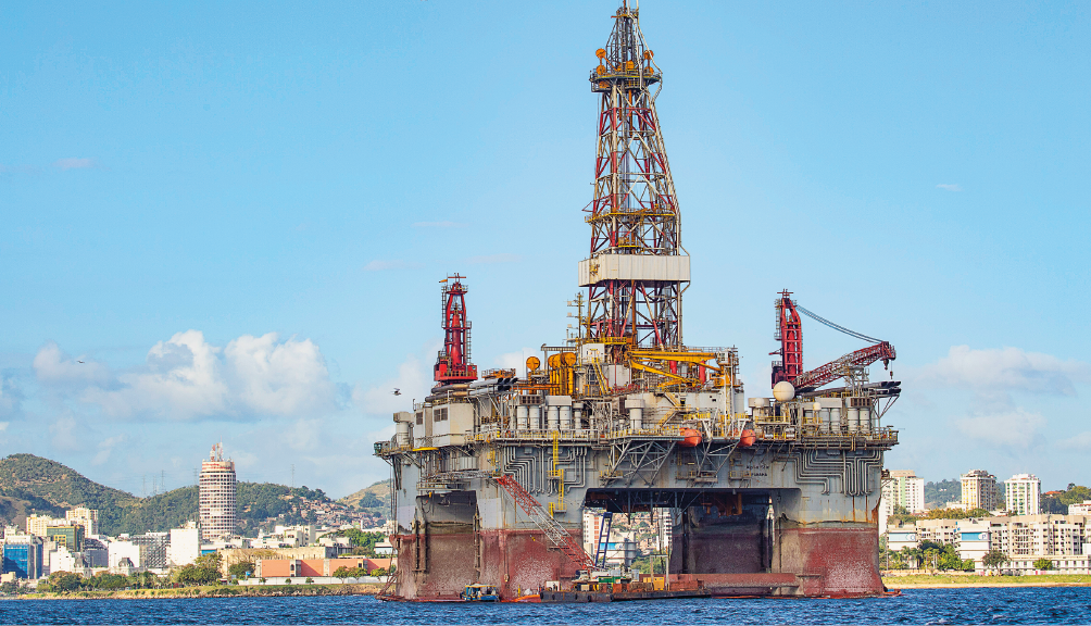 Imagem: Fotografia. Uma plataforma de petróleo no mar. Ao fundo, uma cidade. Fim da imagem.