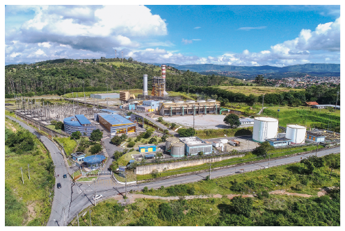 Imagem: Fotografia. Uma fábrica no meio de uma área com vegetação. Há alguns prédios, duas construções cilíndricas e uma torre. Fim da imagem.