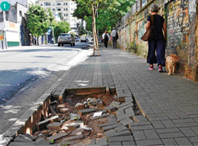 Imagem: Fotografia. Uma mulher de calça jeans e camiseta azul marinho em uma calçada de piso intertravado. Ela segura a coleira de um cachorro bege. Ao lado dela, um buraco na calçada com as peças do piso soltas.  Fim da imagem.