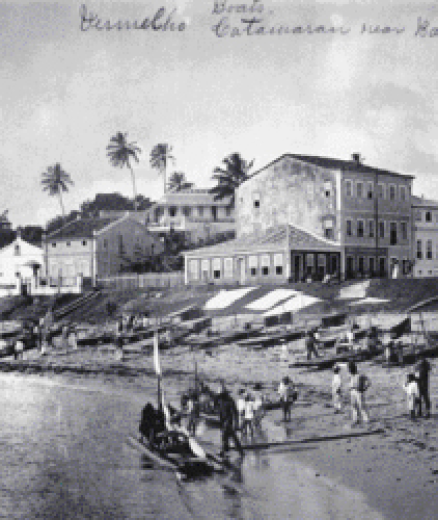 Imagem: Fotografia. Imagem em preto e branco de uma praia com várias canoas de madeira na areia e uma no mar, com pessoas ao redor. No fundo, construções antigas.  Fim da imagem.