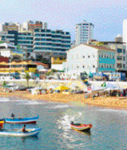 Imagem: Fotografia. Uma praia com canoas coloridas na água. Na areia, tendas com pessoas. Atrás, cidade com prédios e casas.  Fim da imagem.