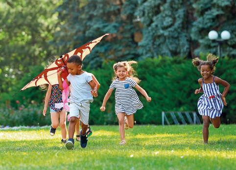 Fotografia 2. Quatro crianças com características diversas correm em uma área verde. Uma delas segura uma pipa.