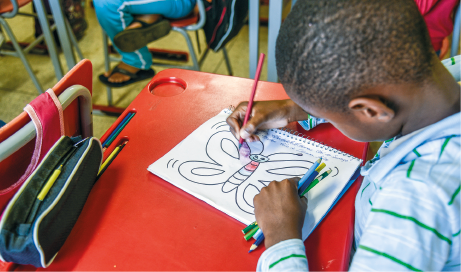 Imagem: Fotografia. Destaque de um menino que pinta o desenho de uma borboleta com lápis colorido. Ele está sentado à carteira, onde também há um estojo.  Fim da imagem.
