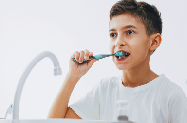Imagem: 3. Fotografia. O menino escova os dentes diante de uma pia. Fim da imagem.