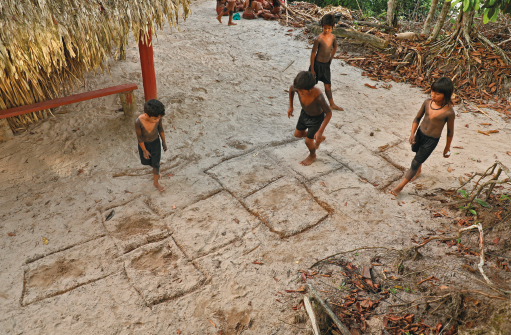 Imagem: Fotografia. Quatro crianças brincam de amarelinha com os quadrados contornados no chão de terra. Uma delas pula enquanto as outras observam.  Fim da imagem.
