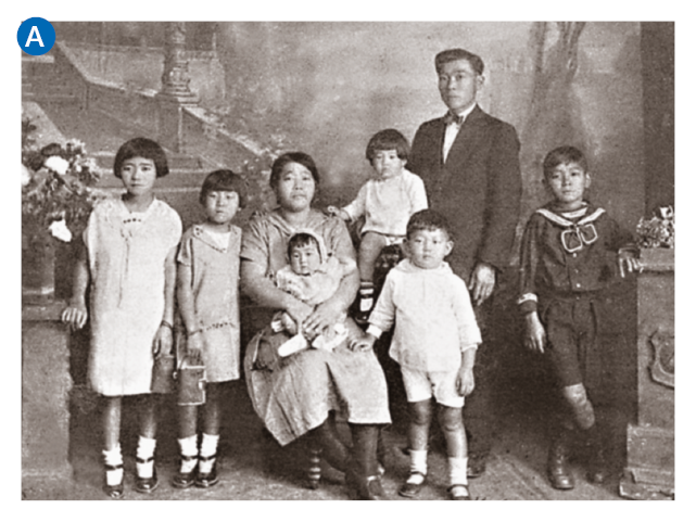 Imagem: A. Fotografia em preto e branco. Uma família que apresenta olhos puxados e cabelo liso escuro posa para foto. É composta quatro crianças grandes, dois meninos e duas meninas, que estão de pé. Uma mulher sentada ao centro com um bebê e ao seu lado, sentada em uma estrutura mais alta, está uma criança pequena. Ao lado dela, um homem de pé que veste terno escuro. Fim da imagem.