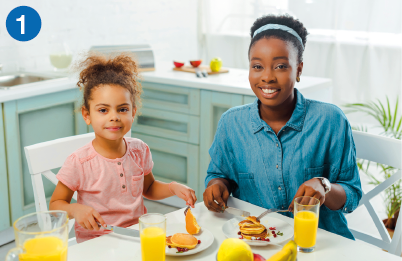 Imagem: Fotografia 1. Fotografia. Uma mulher e uma criança estão lado a lado, sorriem e posam para foto sentadas à mesa posta com suco e frutas. Ao fundo, armários. Fim da imagem.