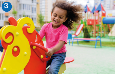 Imagem: 3. A menina sorri e brinca sentada em um cavalo de madeira no parque. Ao fundo, brinquedos.  Fim da imagem.