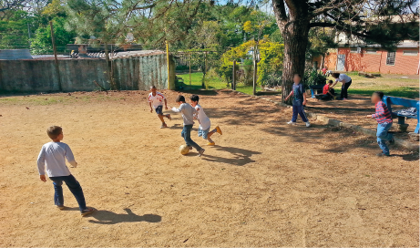 Imagem: Fotografia em plano aberto. Em um terreno, um pequeno grupo de crianças joga bola próximo de uma árvore.  Fim da imagem.
