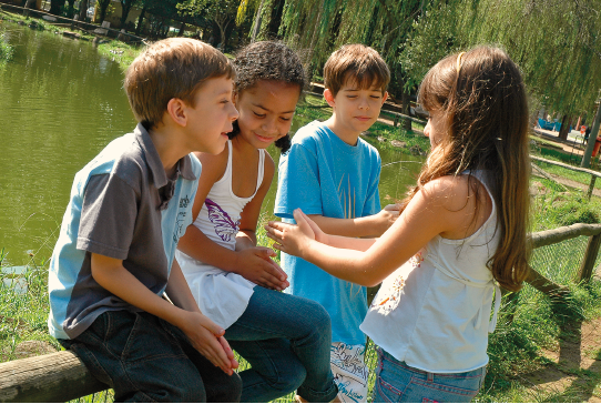 Imagem: Fotografia. Quatro crianças brincam de passa anel em uma área aberta sob o sol e à frente de um lago.  Fim da imagem.