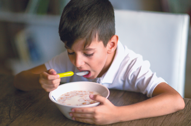 Imagem: 2. Fotografia. O menino está sentado à mesa e faz uma refeição. Fim da imagem.