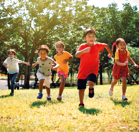 Imagem: Fotografia. Cinco crianças com características diversas sorriem enquanto correm na mesma direção em uma área gramada e sob o sol. Ao fundo, árvores.  Fim da imagem.