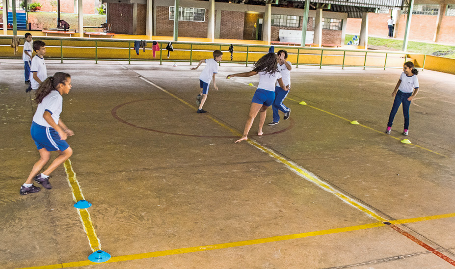 Imagem: Fotografia. Um pequeno grupo de crianças uniformizadas brincam em uma quadra coberto. Elas estão de pé e correm.  Fim da imagem.