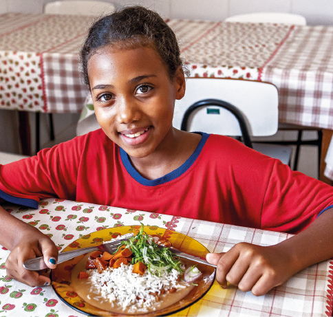 Imagem: Fotografia. Uma menina posa sorridente sentada à mesa diante de um prato de comida. Fim da imagem.