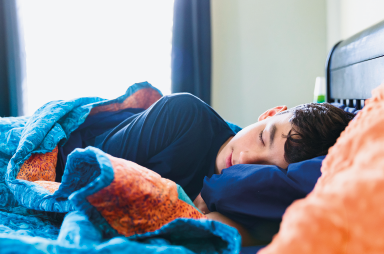 Imagem: 4. Fotografia. O menino está deitado coberto em sua cama.  Fim da imagem.