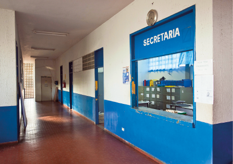 Imagem: Fotografia. Destaque da fachada de uma sala com a identificação “Secretaria”. Há um balcão aberto para o interior do espaço e um amplo corredor do lado de fora.  Fim da imagem.