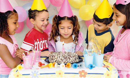 Imagem: Fotografia. Cinco crianças estão diante de uma mesa posta com bolo com uma vela de seis anos, copos coloridos e enfeites. Todas usam chapéus cônicos coloridos e uma menina, que está ao centro, inclina-se ao soprar a vela. Ao fundo, há balões coloridos. Fim da imagem.