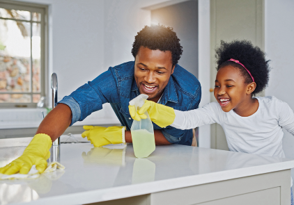 Imagem: Fotografia. Um homem e uma menina sorriem enquanto limpam uma bancada. Ambos estão de luvas. Ele passa um pano na superfície e ela aplica o produto com borrifador.  Fim da imagem.