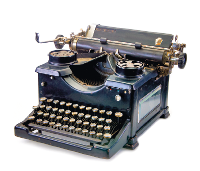 Imagem: Fotografia. Uma máquina de escrever que apresenta muitas teclas redondas e salientes dispostas em quatro fileiras e uma estrutura na parte superior em que há um cilindro horizontalizado ligado a uma pequena manivela e dois círculos, um de cada lado da máquina.  Fim da imagem.