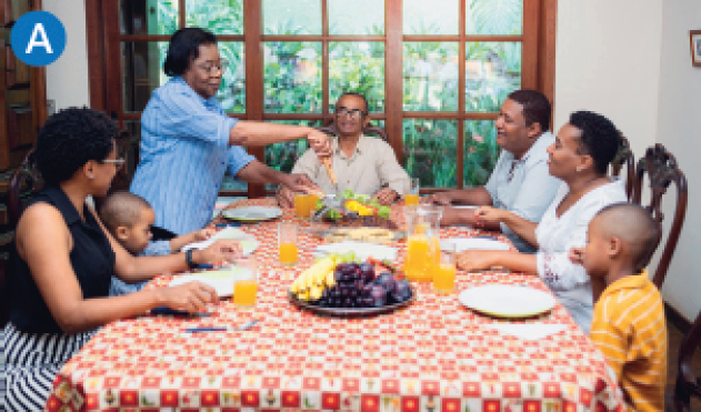 Imagem: Fotografia. Em uma sala ampla, uma família composta por sete pessoas, sendo duas crianças, está reunida à mesa posta com suco, frutas e pratos. Todos estão sentados, com exceção de uma mulher de pé que manipula um dos alimentos.  Fim da imagem.