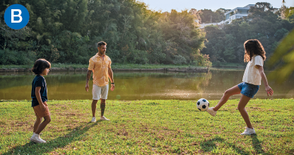 Imagem: B. Fotografia. Um homem adulto e duas crianças, um menino e uma menina, brincam de bola numa área gramada sob o sol. Ambos observam a menina que está com a bola no pé.  Fim da imagem.