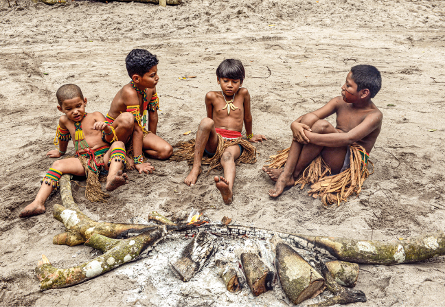 Imagem: Fotografia. Quatro crianças indígenas, de pele escura e cabelo preto, estão sentadas lado a lado na areia próximo de lenhas de uma fogueira apagada. Elas estão de sunga e tanga de palha e usam adereços coloridos no pescoço, nos braços e pernas. Fim da imagem.