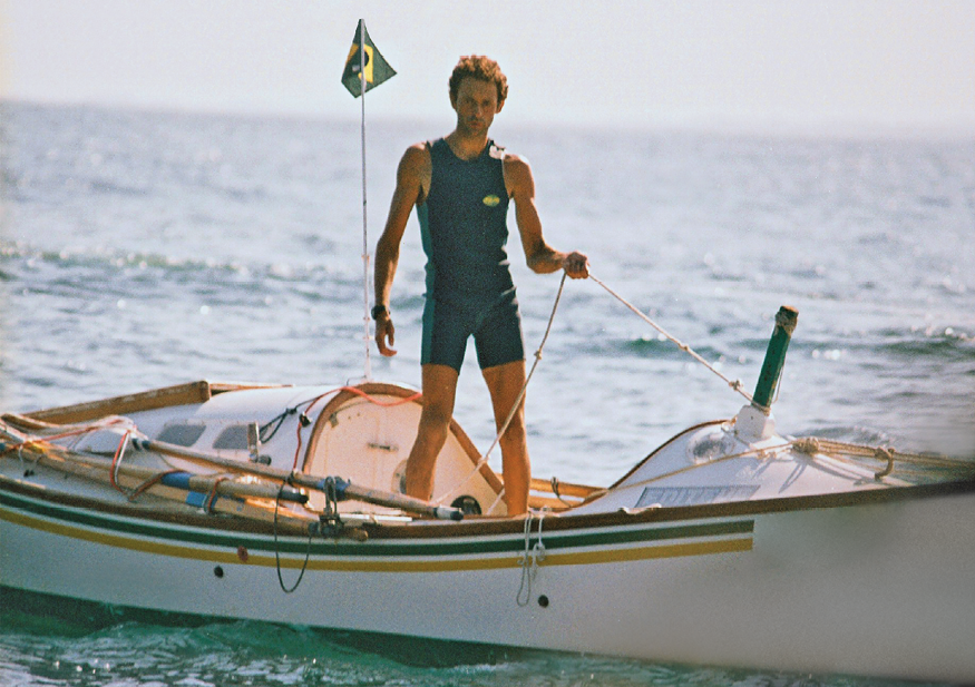 Imagem: Fotografia. Em alto-mar, destaque de um homem de pé em um barco e que veste uma roupa fresca com camiseta regata e shorts. Ele segura uma corda acoplada à embarcação e olha para foto. Ao fundo, há uma pequena bandeira do Brasil hasteada.  Fim da imagem.