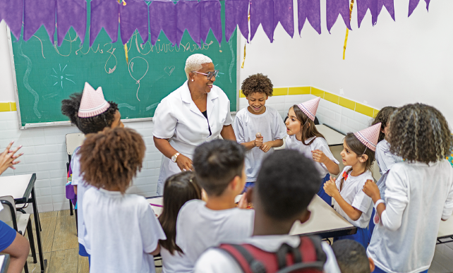 Imagem: Fotografia. Em uma sala de aula enfeitada com bandeirolas roxas penduradas em um varal, um grupo de alunos uniformizados, alguns usando chapéu cônico, e a professora de jaleco sorriem e alguns batem palma. Na lousa, está escrito: “Feliz Aniversário” e a figura de um balão.  Fim da imagem.