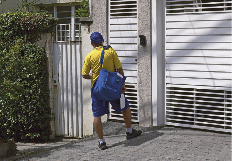 Imagem: Fotografia. Um homem uniformizado com boné azul, camiseta amarela, bolsa larga azul transpassada no ombro, bermuda azul segura envelopes e está parado diante do portão de uma casa.  Fim da imagem.