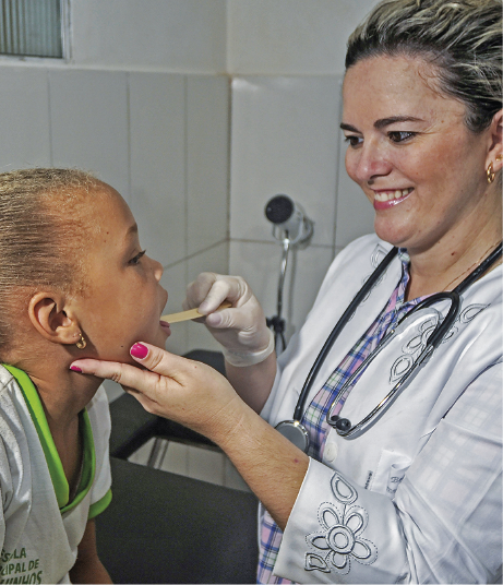 Imagem: Fotografia. Uma mulher de jaleco e com estetoscópio no pescoço sorri enquanto avalia a boca de uma criança com uma pequena espátula.  Fim da imagem.