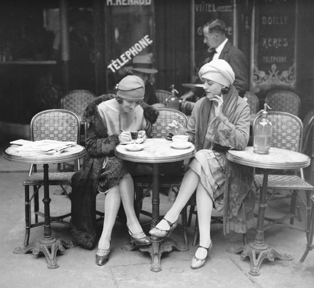 Imagem: Fotografia em preto e branco. Duas mulheres de pele clara estão sentadas diante de uma pequena mesa redonda de um café. Ambas manipulam xícaras, usam um chapéu pequeno de tecido sobre o cabelo curto, usam vestidos até a altura dos joelhos, casacos longos, meias e sapatos baixos.  Ao redor, outras pequenas mesas com cadeiras, um garçom ao fundo e uma cabine telefônica na qual está escrito: “Télephone”.  Fim da imagem.