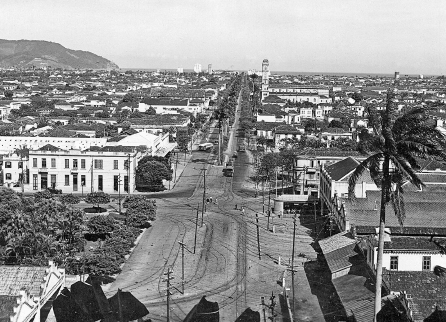 Imagem: Fotografia em preto e branco. Em plano aberto, destaque de uma via extensa rodeada por amplas construções assobradas e árvores.  Fim da imagem.