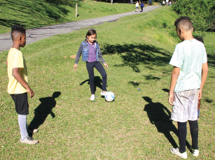 Imagem: Fotografia. Três crianças estão de pé e brincam de bola em uma área gramada sob o sol. Fim da imagem.
