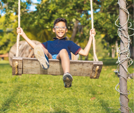 Imagem: Fotografia. Um menino de óculos sorri sentado em um balanço de madeira e corda em uma área verde.  Fim da imagem.