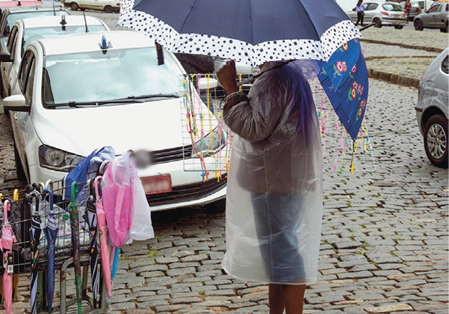 Imagem: Fotografia. Destaque de uma pessoa com capa de chuva e sombrinha que está parada em uma via de paralelepípedos enquanto vende guarda-chuvas expostos próximo à guia da calçada. Ao fundo, carros.  Fim da imagem.
