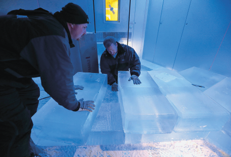 Imagem: Fotografia. Dois homens agasalhados e de luvas, um deles de toca, estão dentro de um espaço fechado com luz azulada e manipulam grandes barras de gelo.  Fim da imagem.